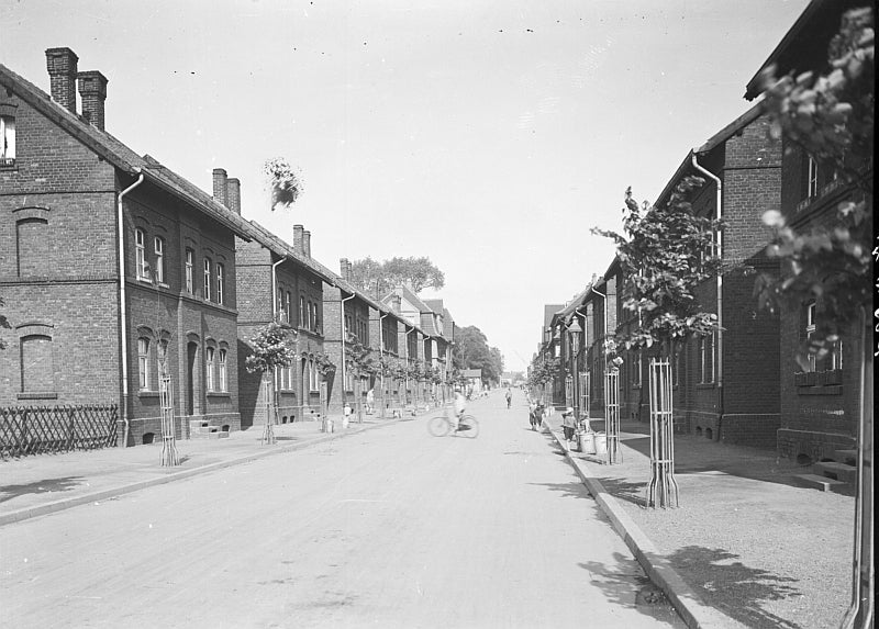 Altes schwarz-weiß Foto einer Siedlung mit Häuser. Auf der Straße gehen Menschen und eine Person überquert die Straße mit einem Fahrrad.