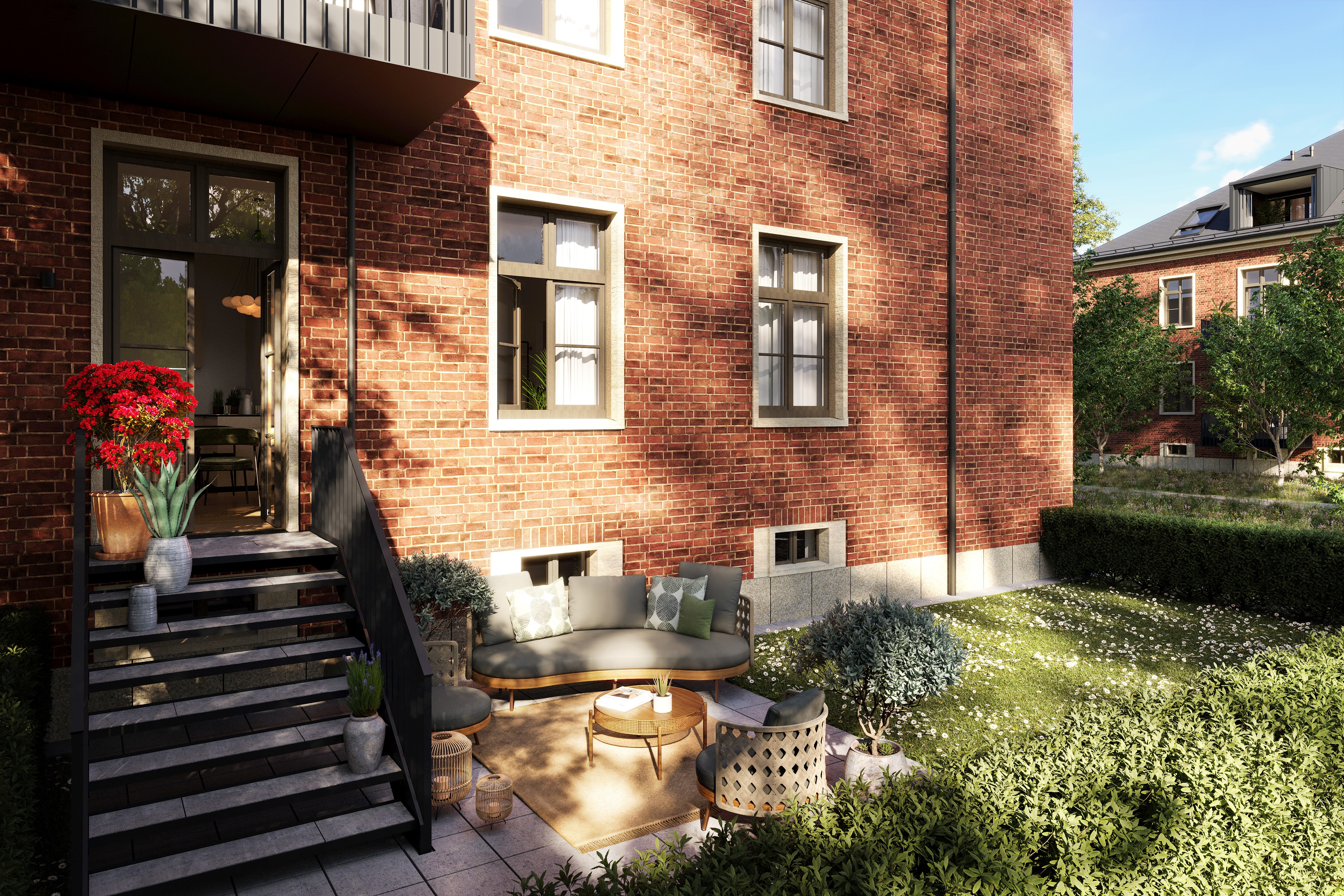 You can see a terrace on the ground floor with stairs to the flat entrance. On the terrace is a couch with carpet, coffee table and chairs. There are flowers on the stairs.