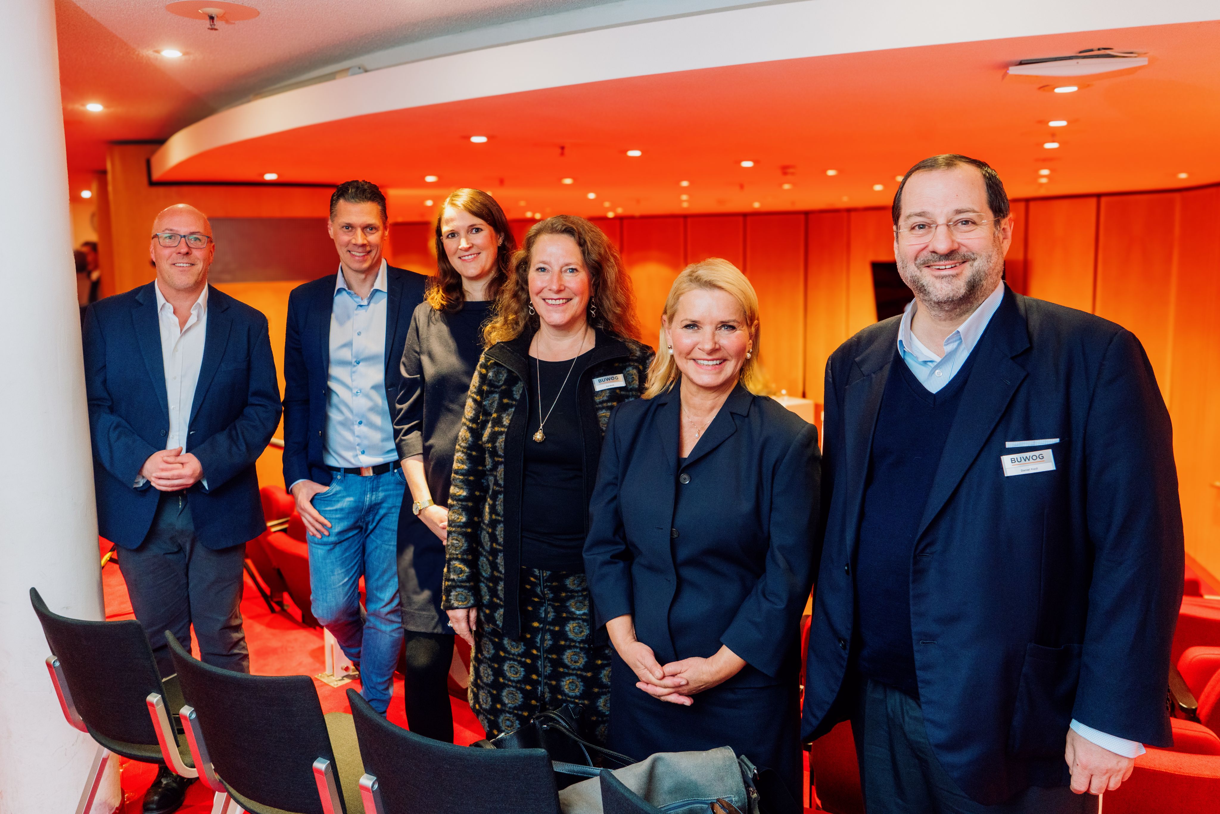 Zu sehen sind 6 Personen, 3 Männer und 3 Frauen mit einem rot-orangenen Hintergrund.